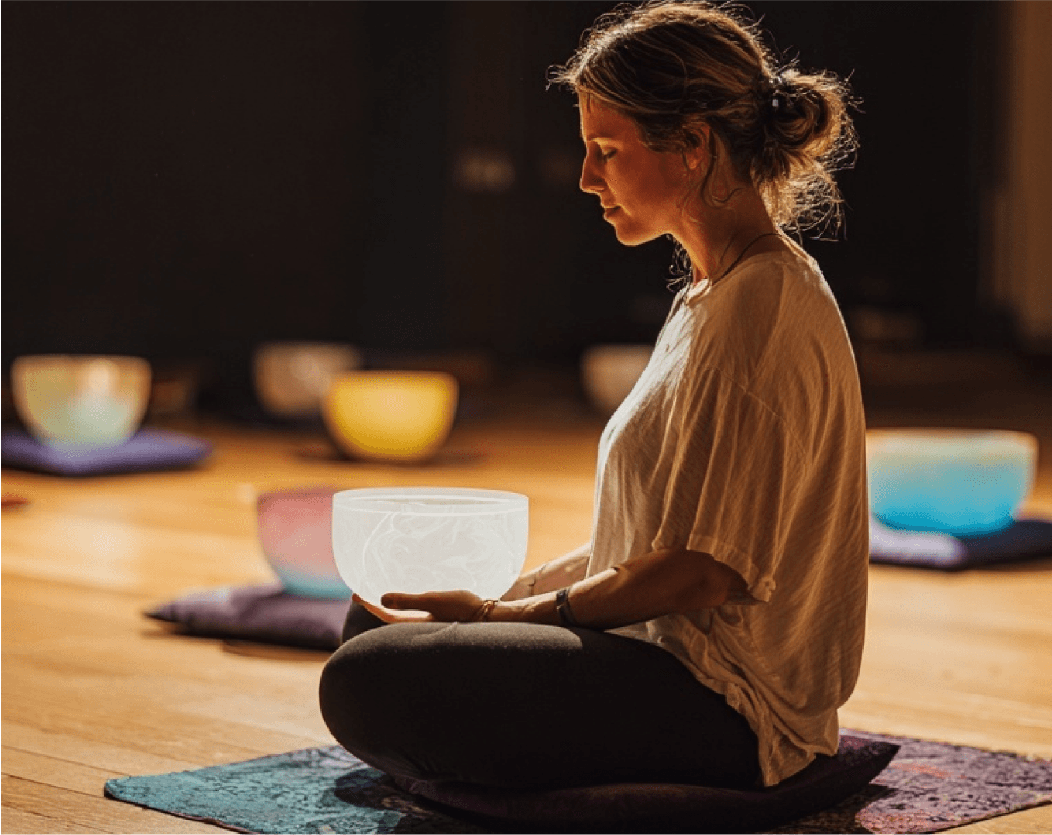 A woman meditating with a frosted crystal singing bowl in her lap, more bowls on cushions behind her.